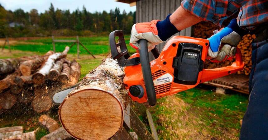 Operator in gloves cutting a log with an orange petrol chainsaw beside a woodpile