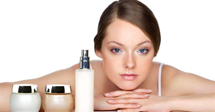 Woman resting beside jars of face cream and a bottle of lotion on a white surface