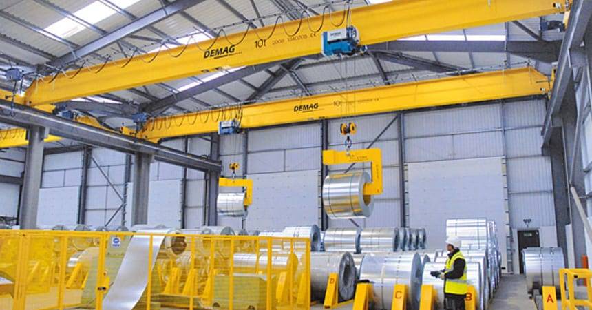 Yellow overhead travelling cranes lifting steel coils inside a warehouse