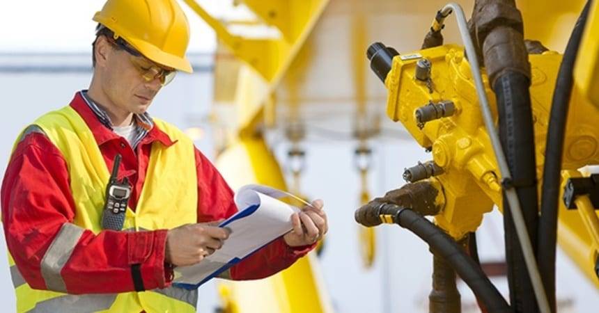 Inspector in a yellow hard hat writing on a clipboard beside yellow industrial machinery