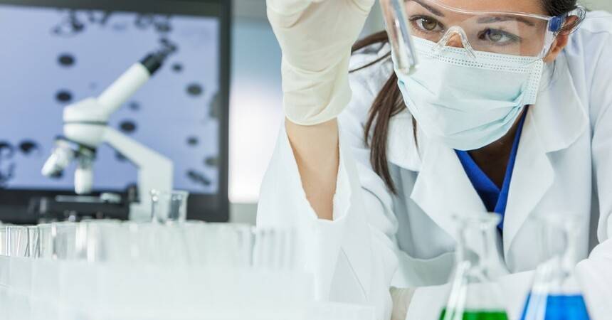 Laboratory scientist in mask and goggles examining a test tube of clear solution