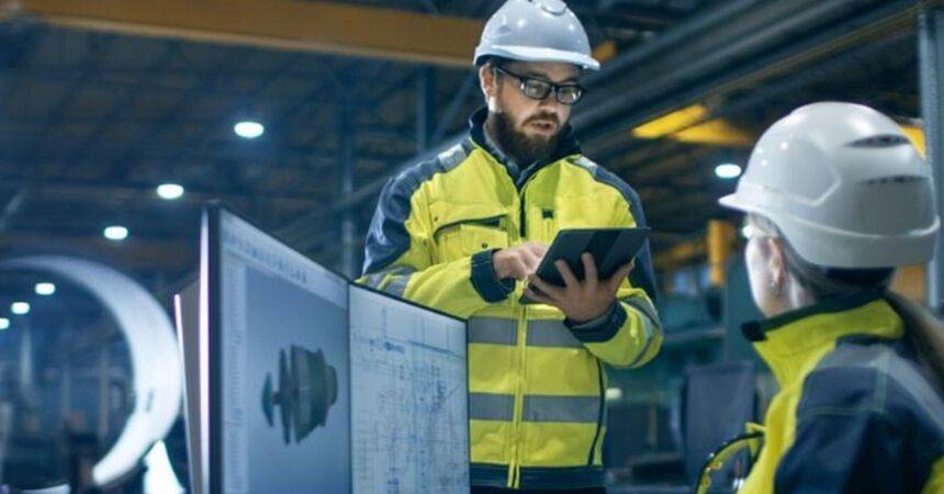 Two engineers in hard hats and high-visibility jackets reviewing drawings on a tablet inside a factory